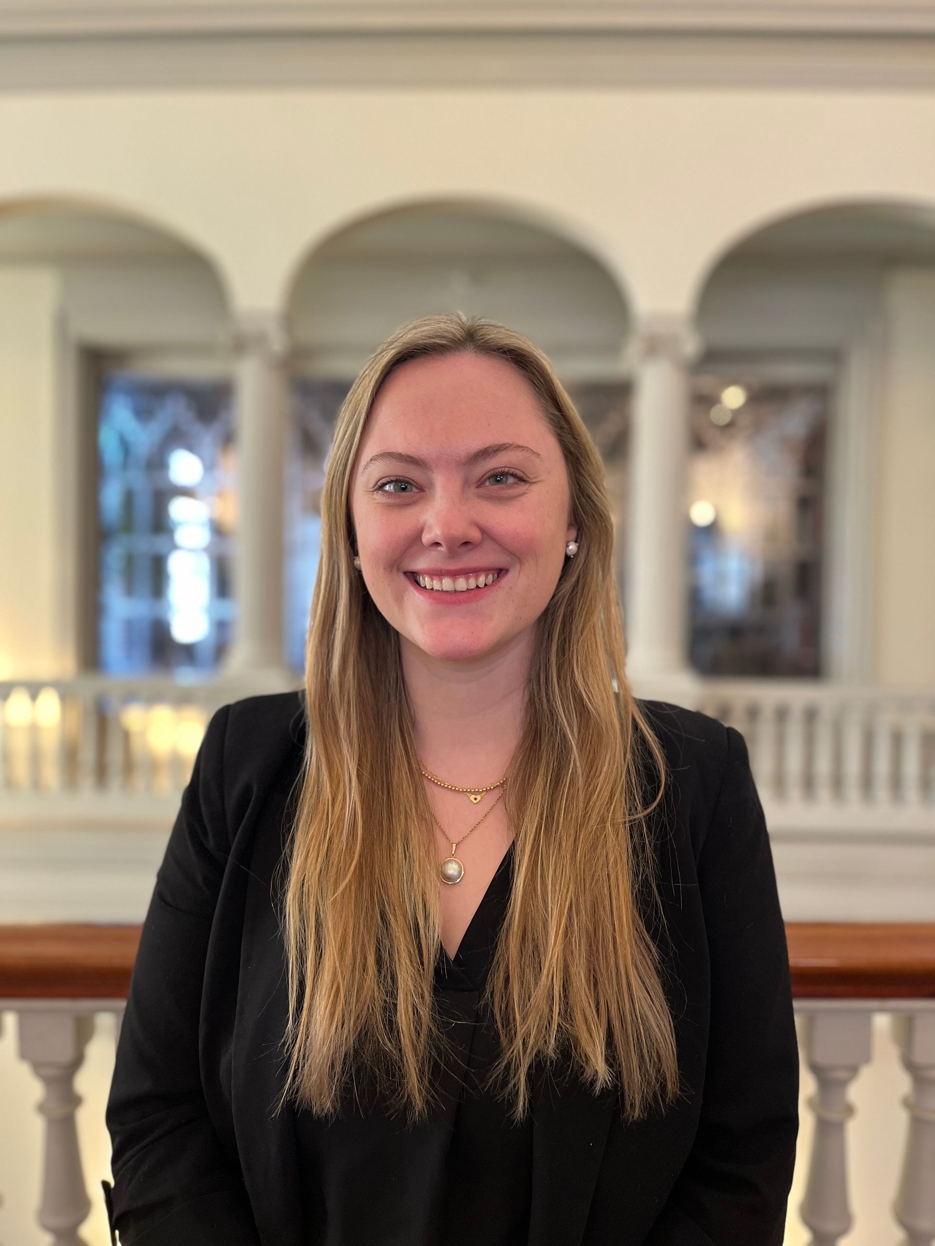 Woman with blonde hair smiling, wearing a black blazer. Interior with columns and arches in background.