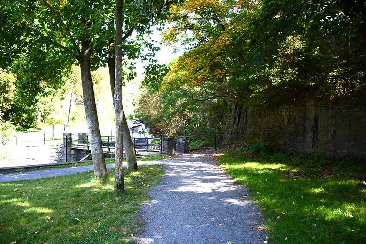 Pathway alongside a waterway and stone wall, bordered by trees and grass.