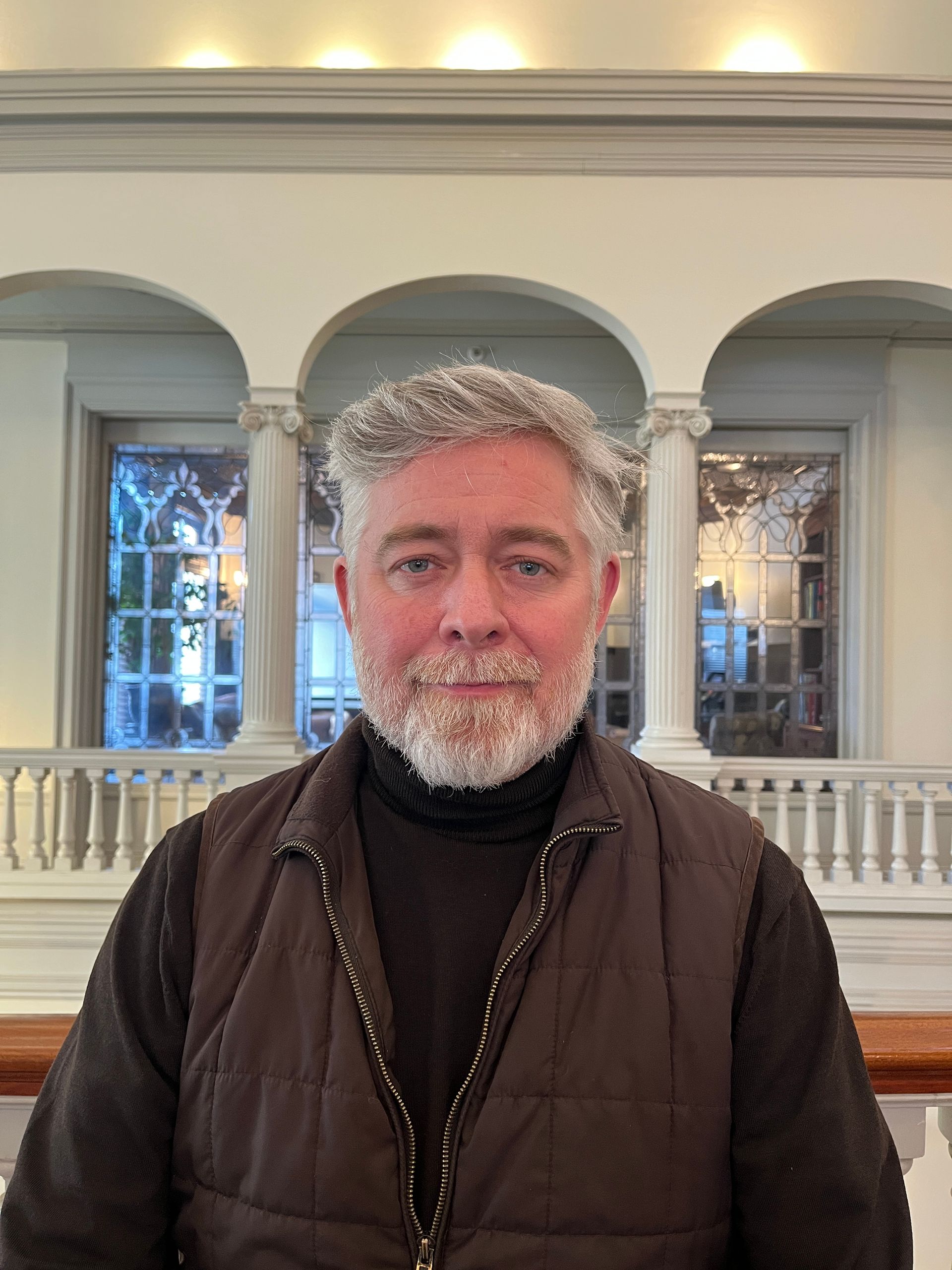 Man with salt and pepper beard in black turtleneck and brown vest, standing indoors with arched windows.