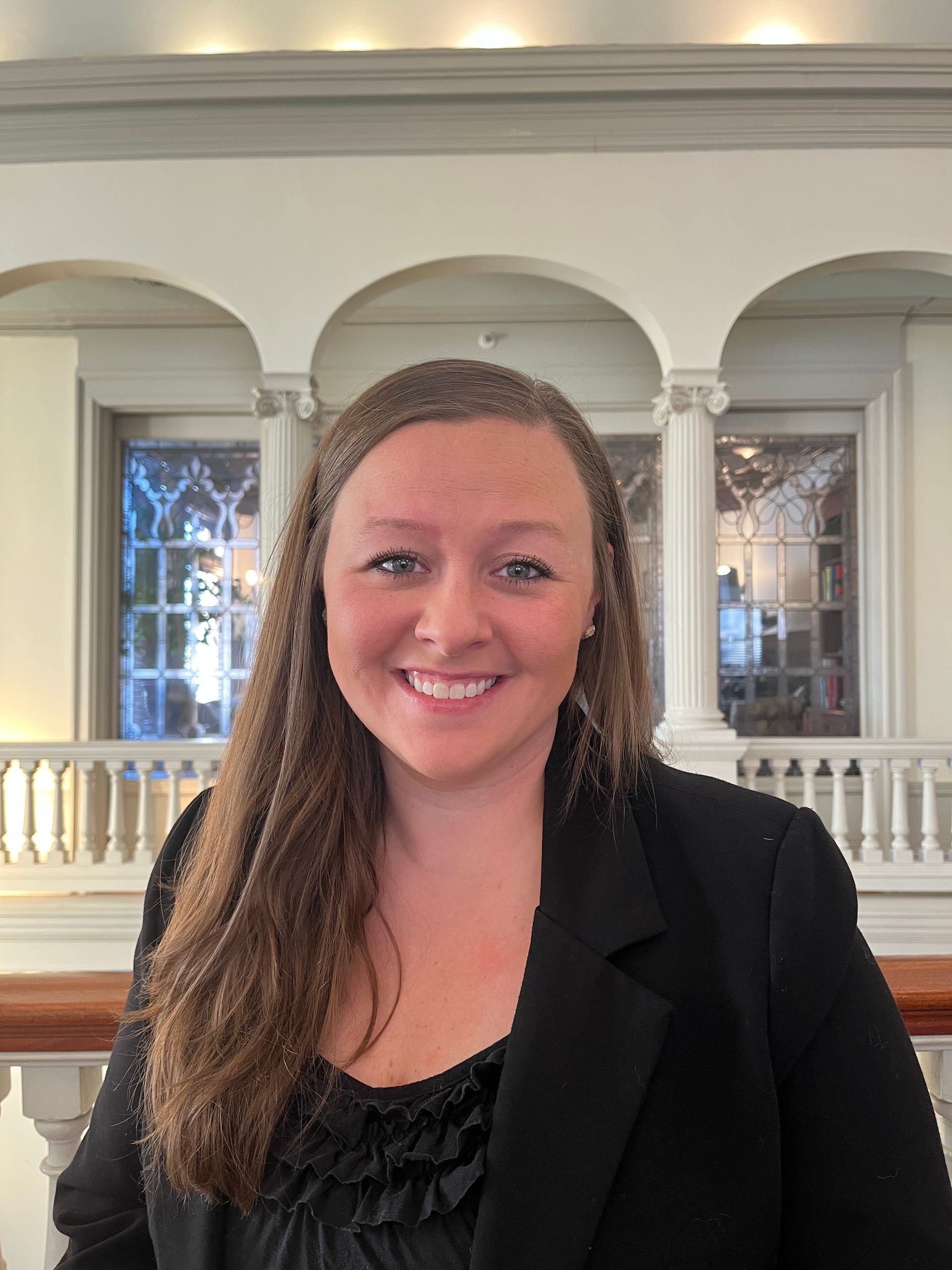 Woman smiles, wearing black blazer. Standing in hall with white columns, and windows in background.