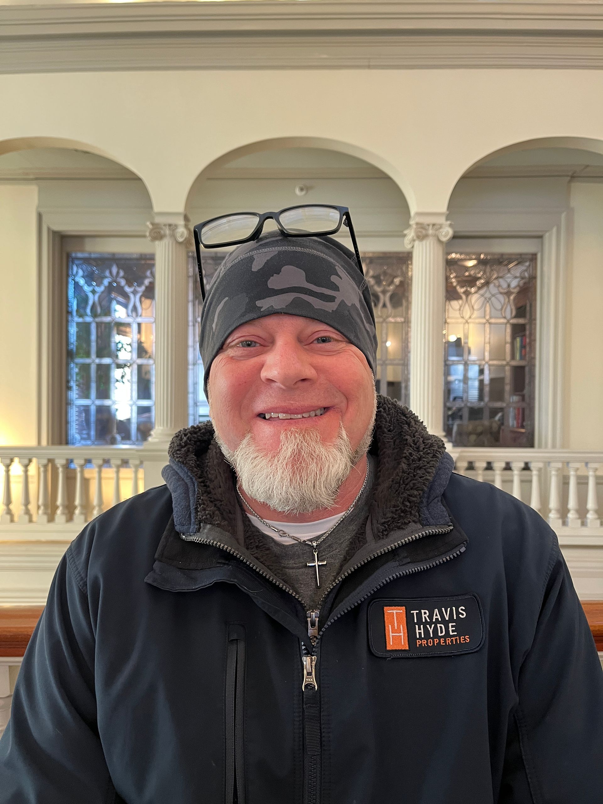 Man wearing a beanie and glasses on top, smiling. Standing inside near arches and windows.