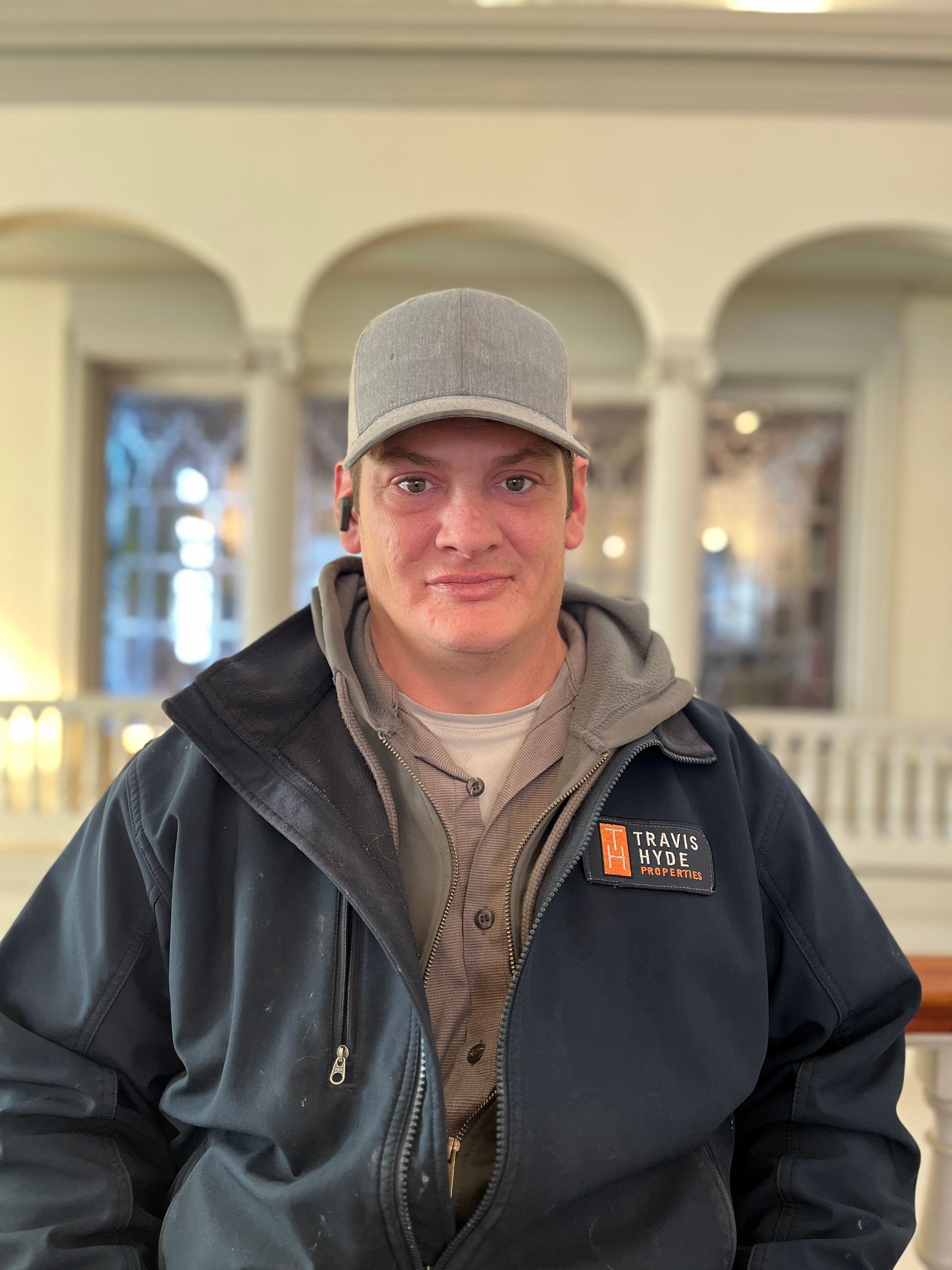 Man in work attire with a gray cap smiles inside a building with arches and pillars.
