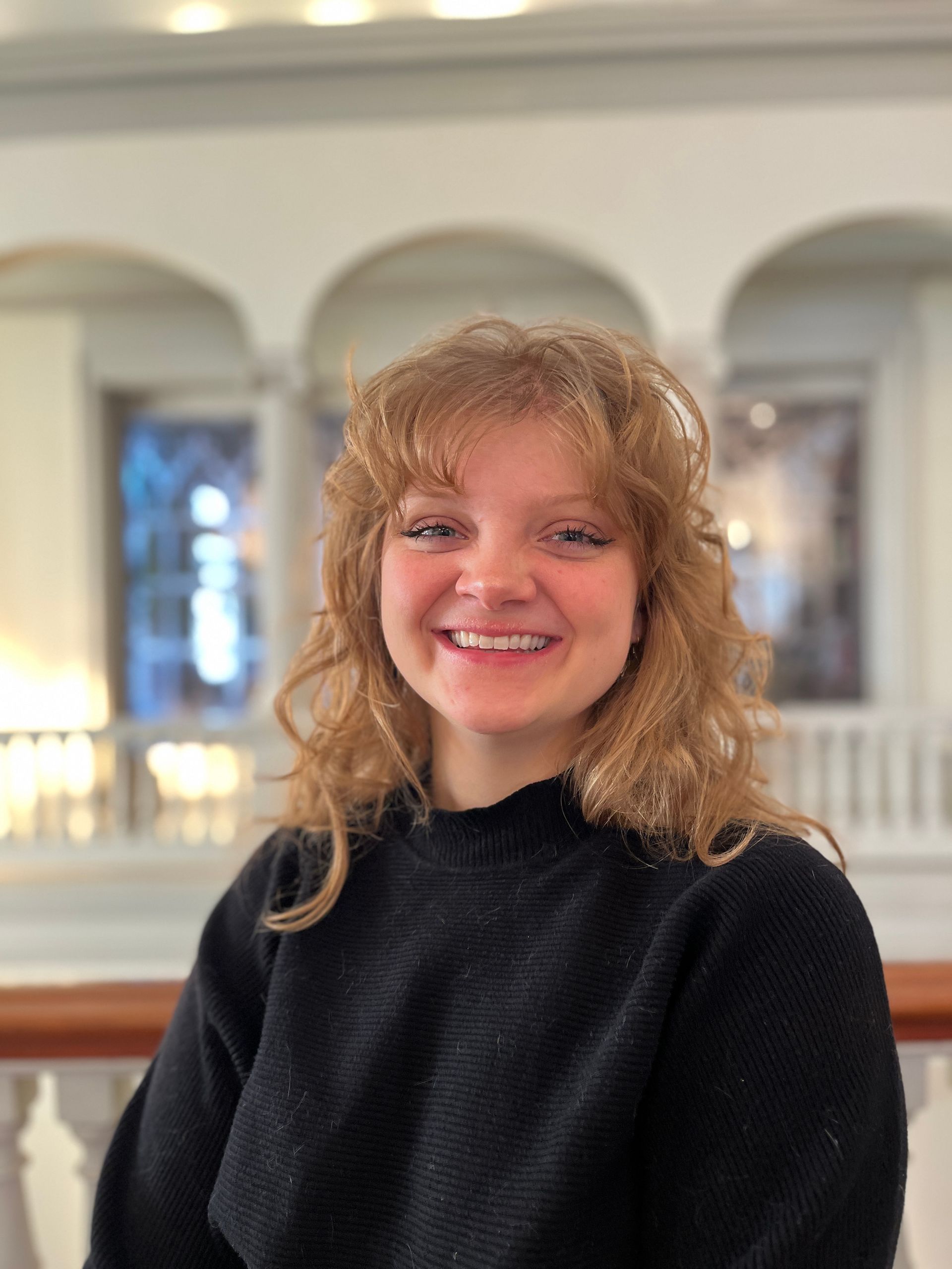 Woman with curly red-blonde hair, smiling with braces, wearing a black sweater, standing in a building with archways.