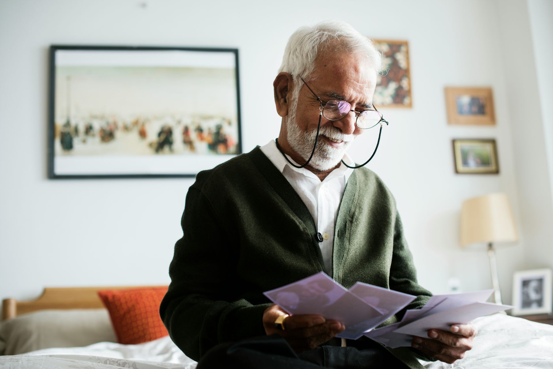 An elderly man is sitting on a bed reading a letter.