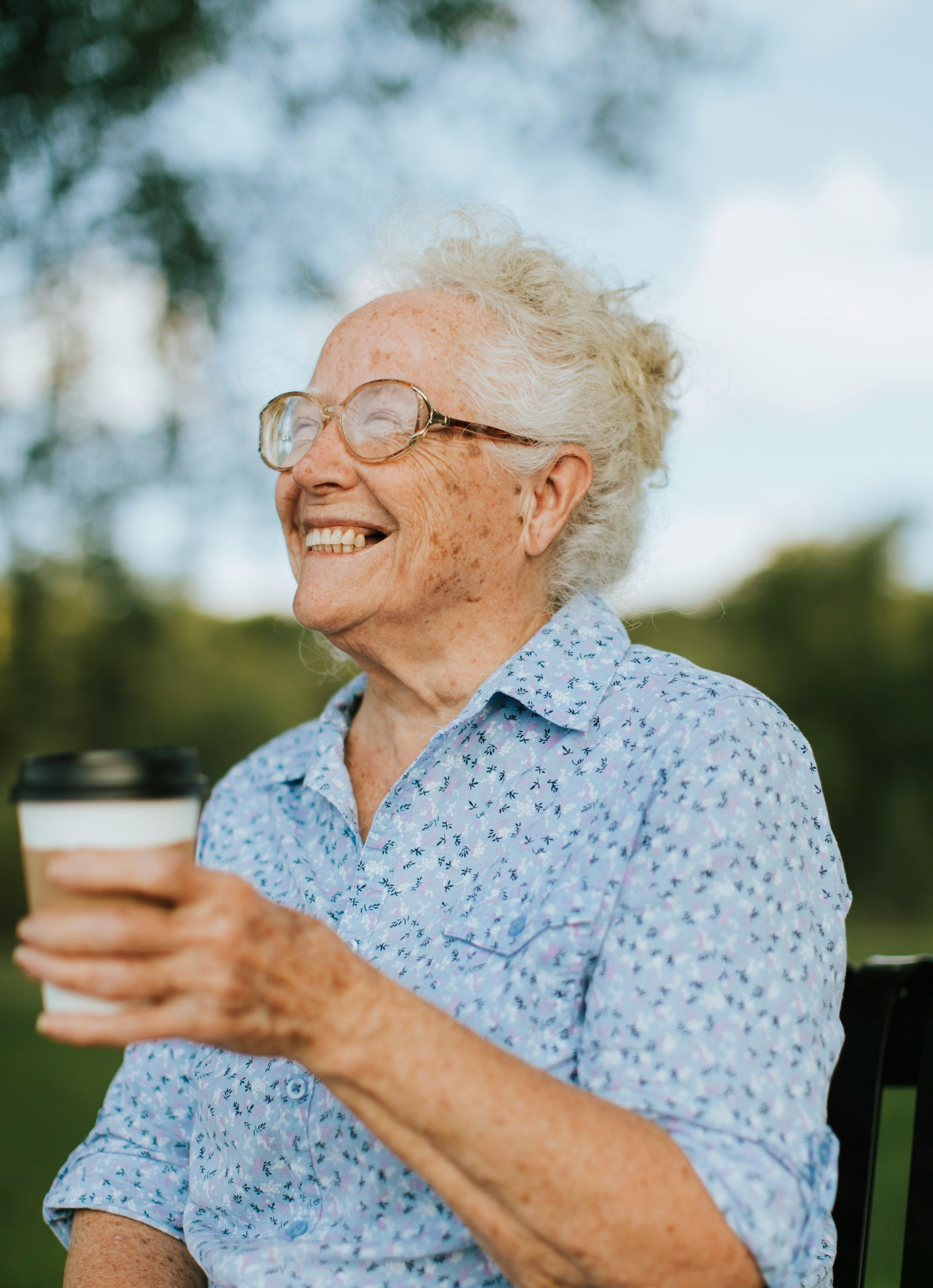 An elderly woman is sitting on a bench holding a cup of coffee and smiling.