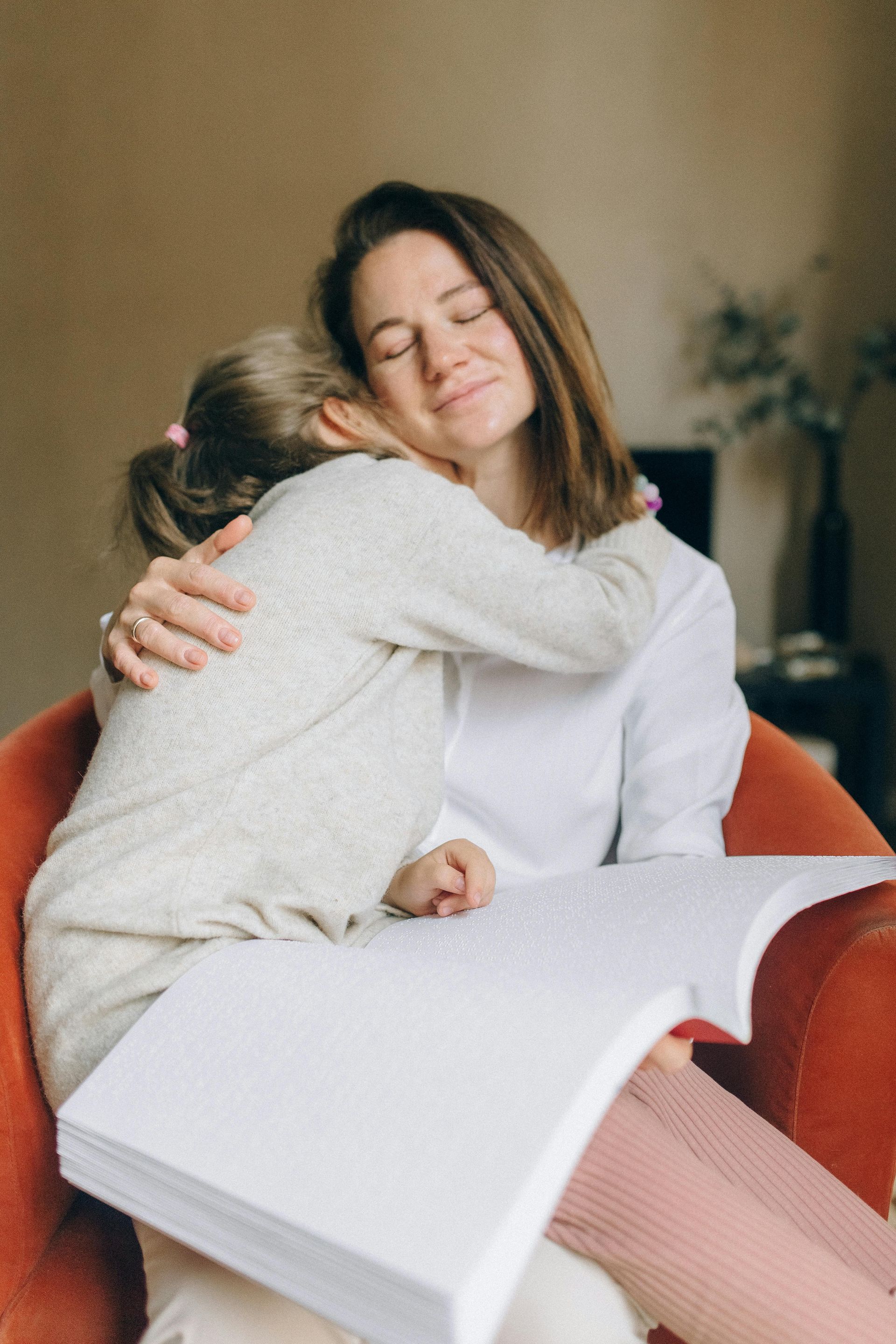 A woman is sitting in a chair hugging a little girl who is reading a book.