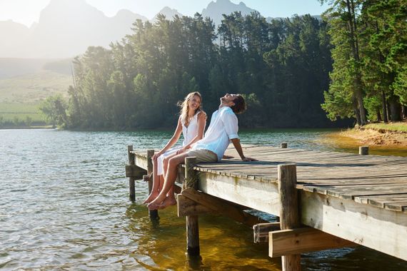 Two people sit on a wooden dock by a lake, feet in the water, with trees and hills in the background.