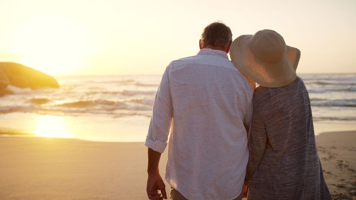 Two people in hats walk hand in hand on a beach at sunset.