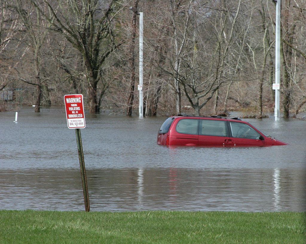 A flooded parking lot with a red minivan. Water nearly up to the top of the car.