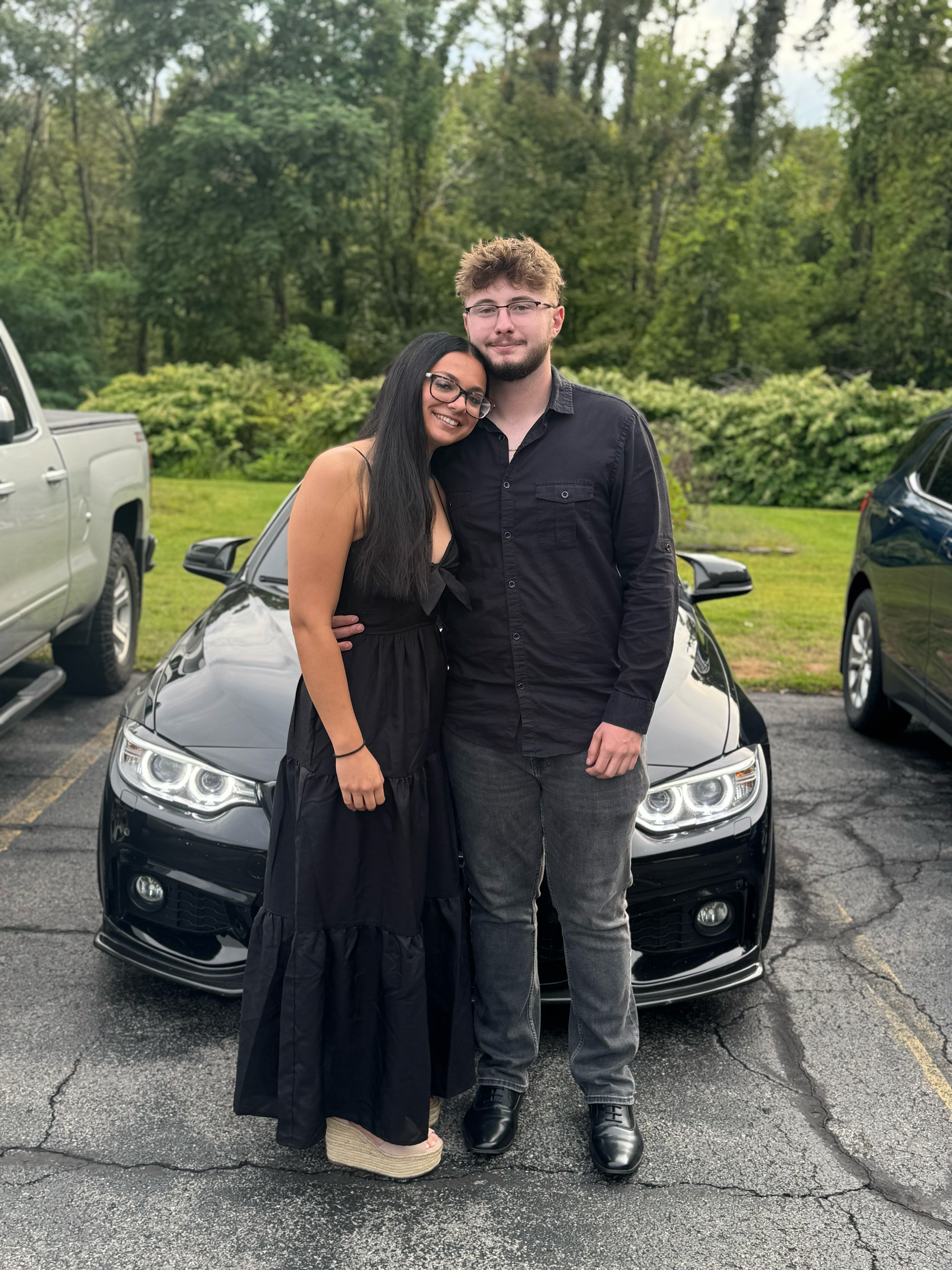 A couple stands together smiling in a parking lot in front of a dark sedan, both wearing black clothing.