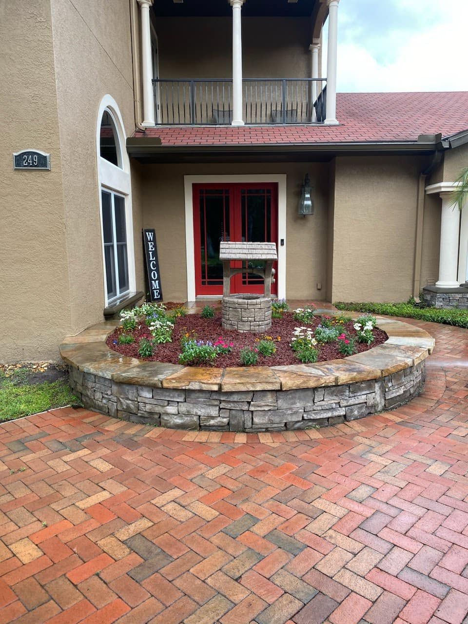 A stone-walled circular garden bed with a small wishing well centerpiece in front of a house with a red front door.