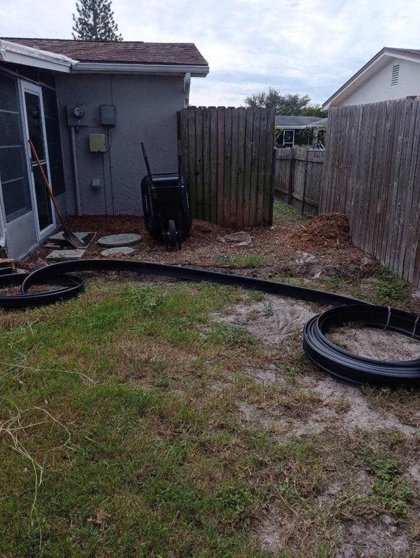 A black landscape edging strip is laid out in a curve on a patchy lawn next to a house and a wooden fence.