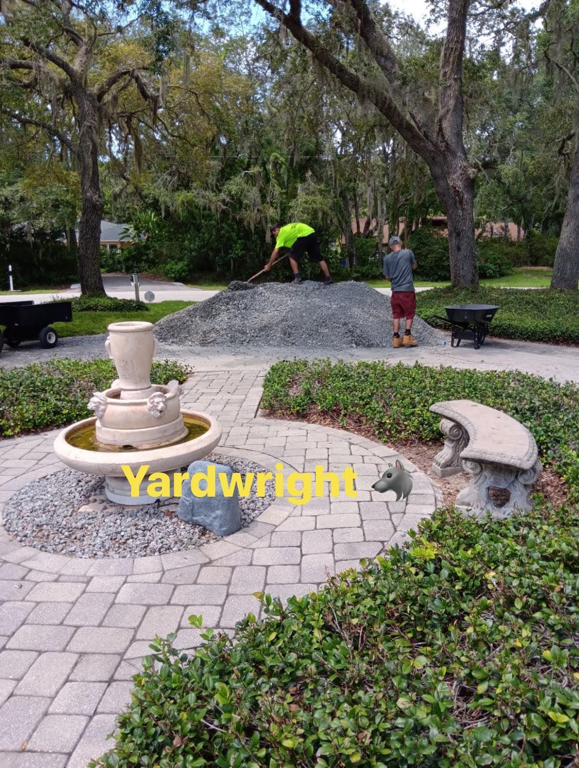 Two workers shovel a large pile of gravel near a stone fountain and garden bench on a paved patio.