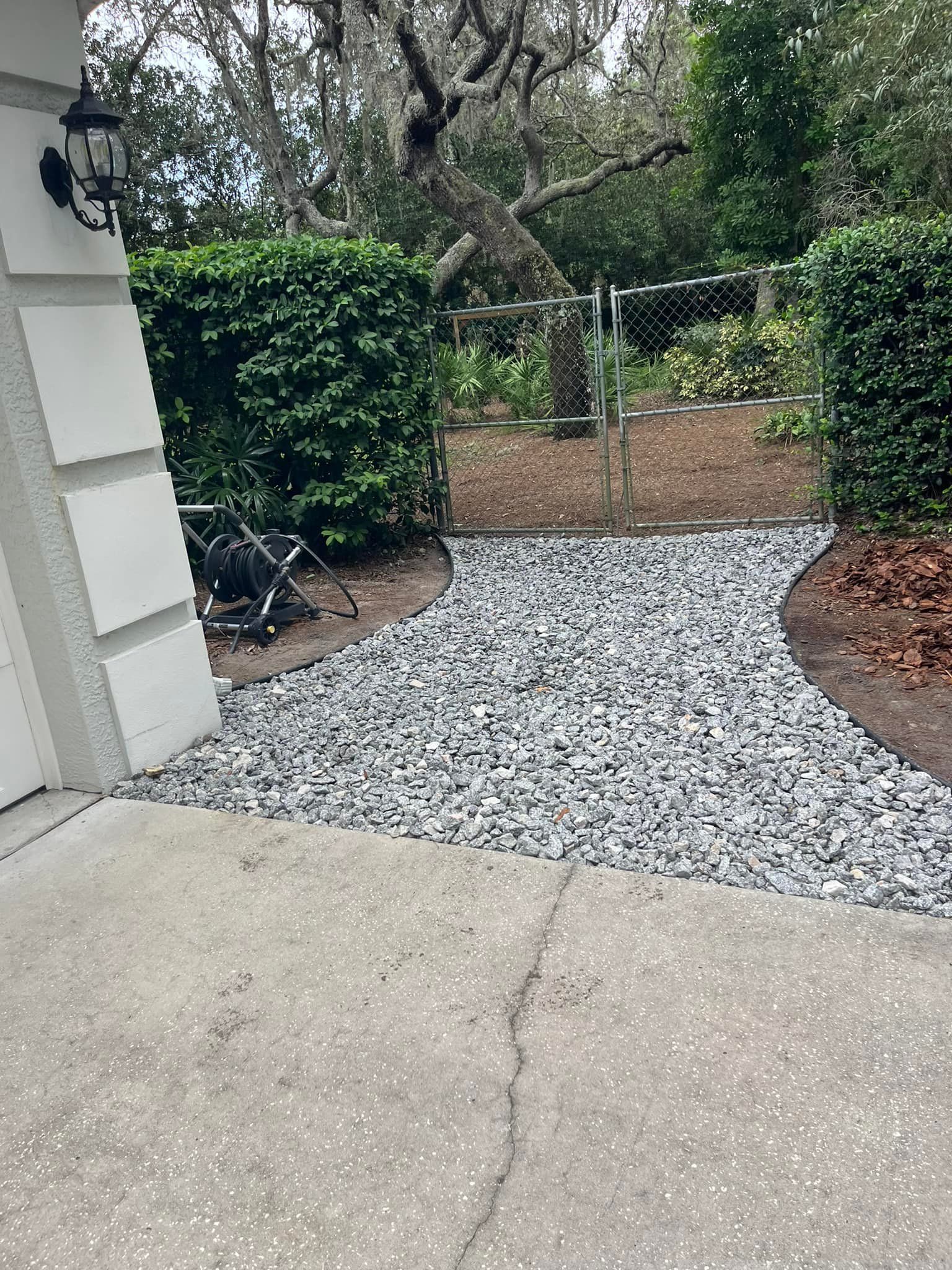 A gray gravel path leads from a concrete driveway to a chain-link gate flanked by hedges.
