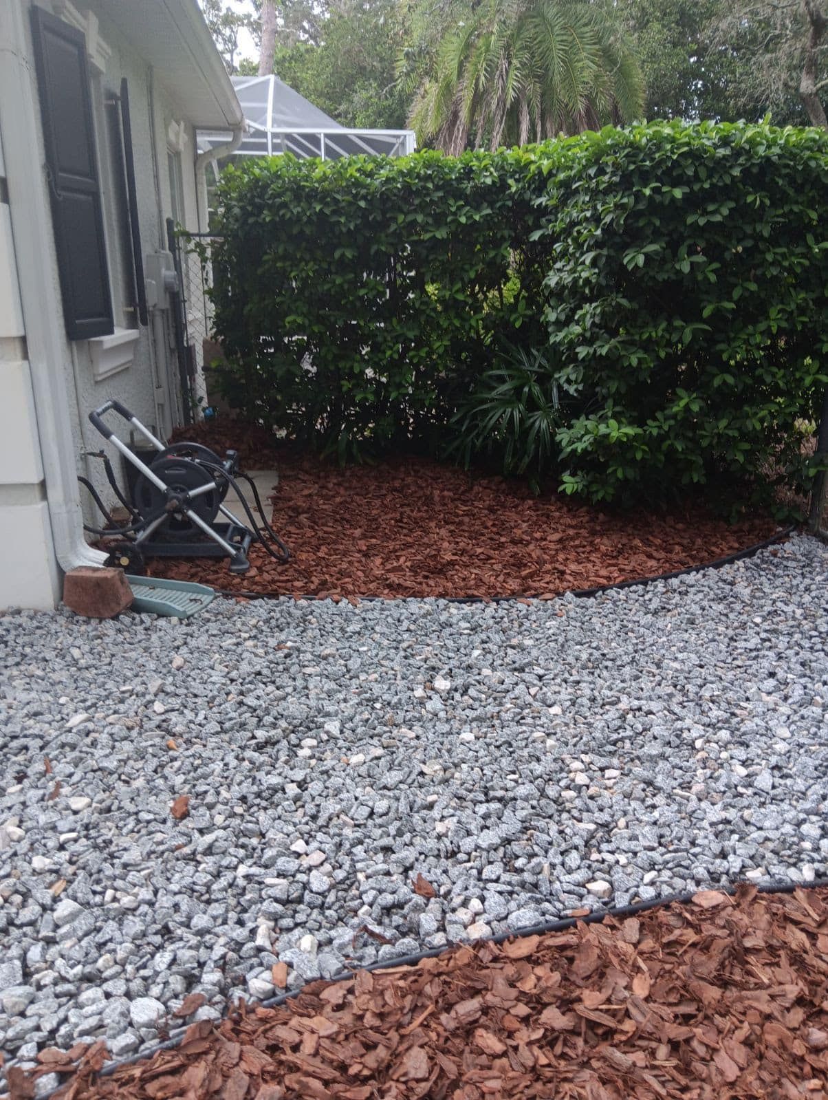 A residential yard with a grey stone pathway separating two sections of fresh reddish-brown mulch next to a house wall.