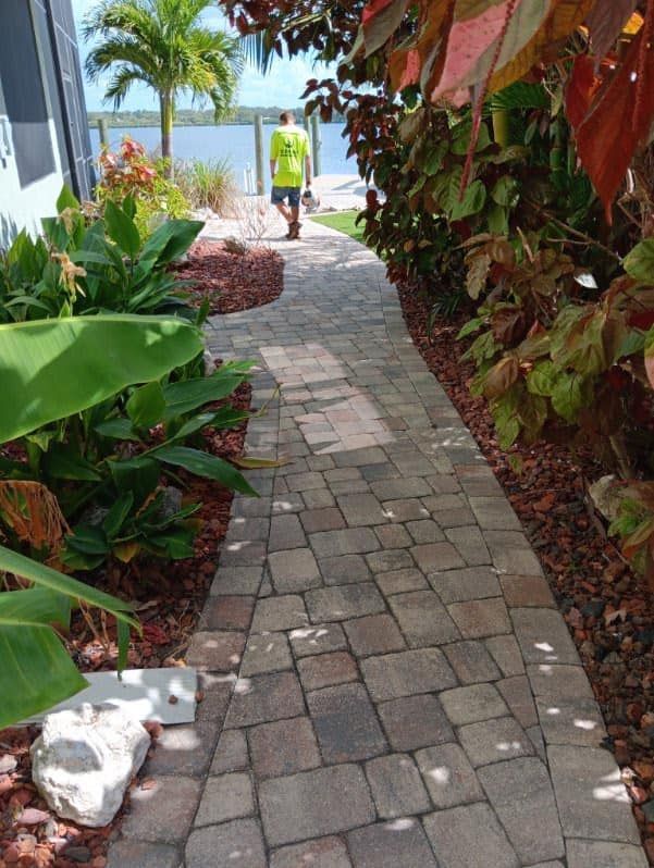 A paved walkway leads toward a person walking toward a body of water, flanked by tropical plants and reddish mulch.