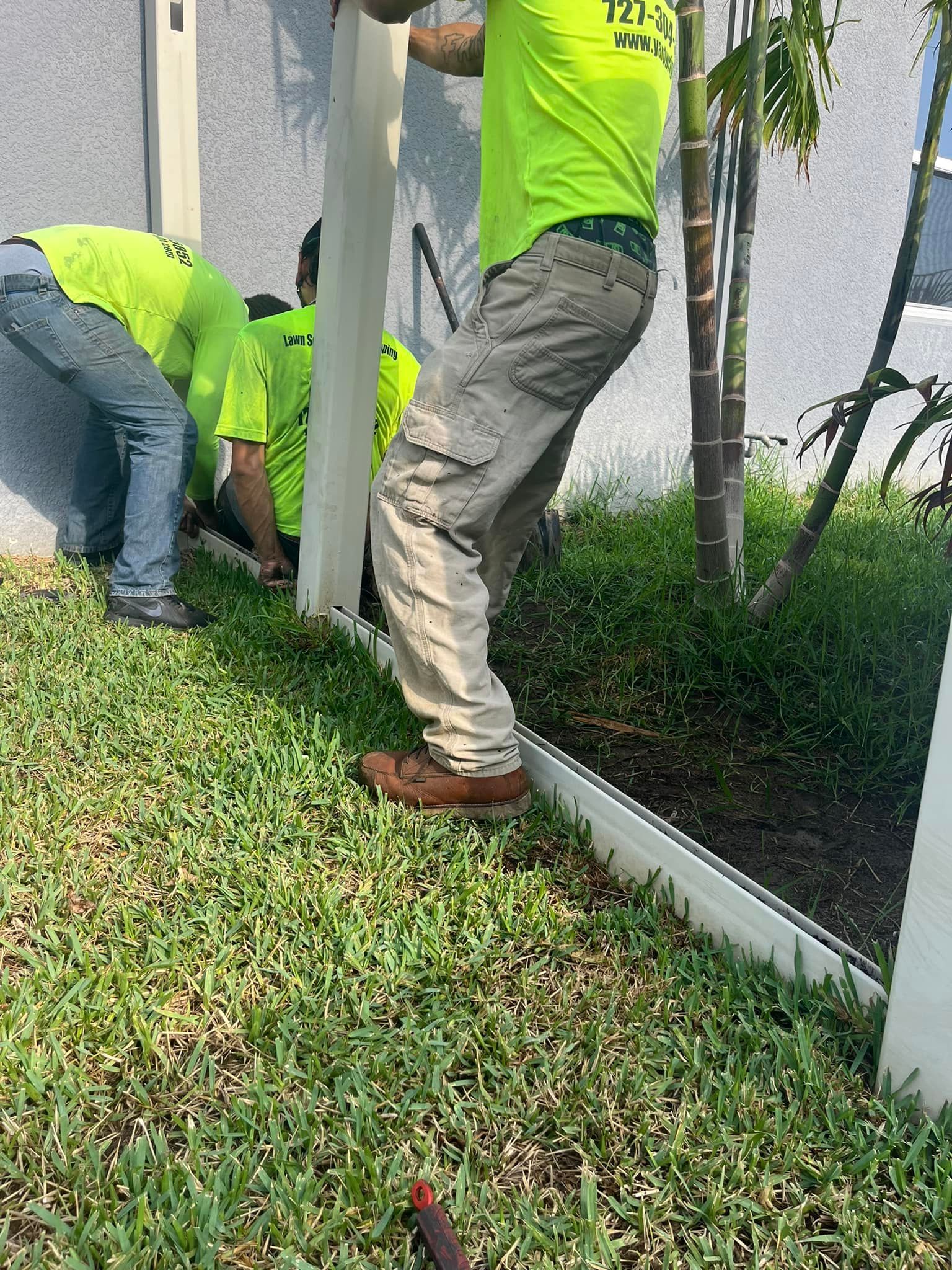 Two workers in neon-yellow shirts install a white fence post into the grass against a light gray wall.