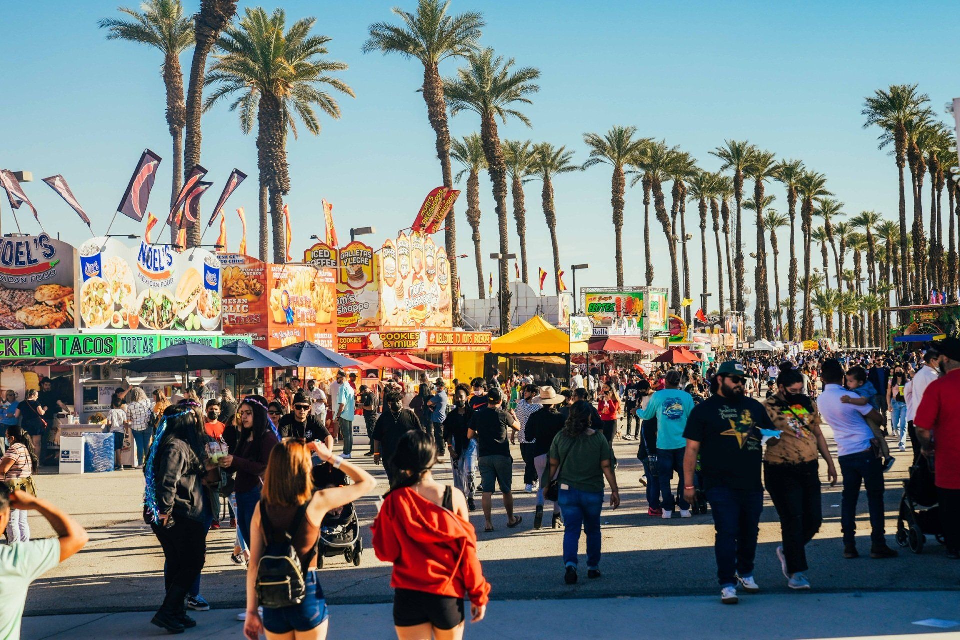 A crowd of people are walking around a carnival with palm trees in the background.