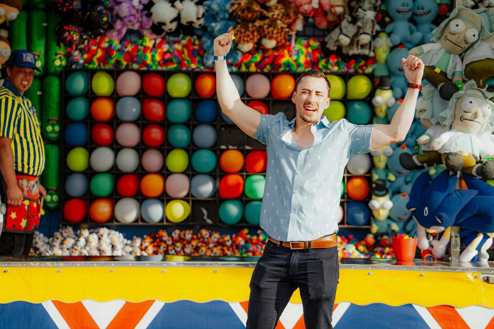 A man is standing in front of a carnival game with his arms in the air.