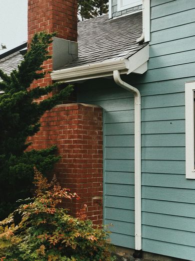 Brick chimney next to a blue-sided house, with white gutters, and green bushes.