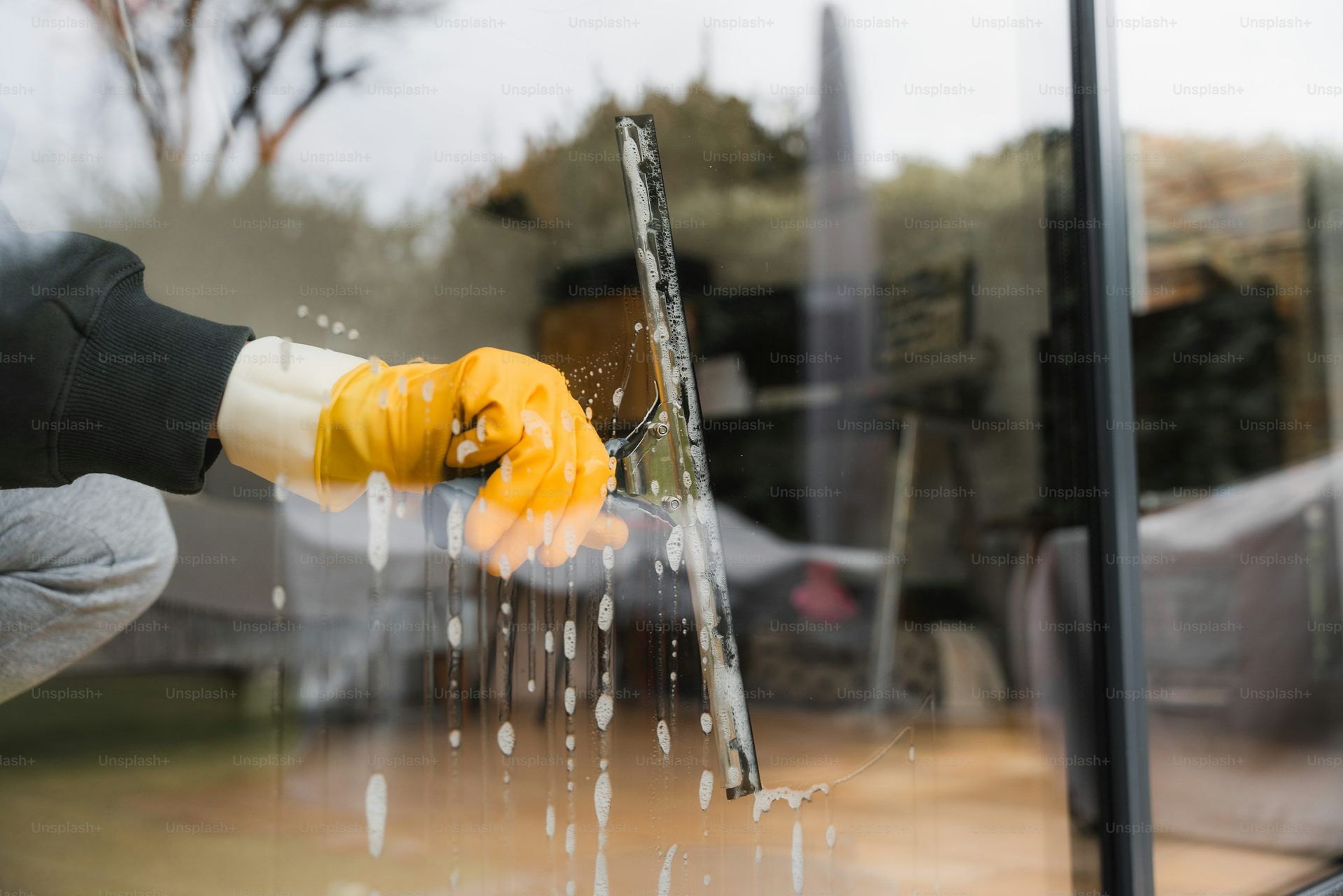 Person wearing yellow gloves cleaning a window with a squeegee; soapy water drips down.