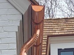 Copper downspout and gutter on the side of a house near a shingled roof and bare trees.
