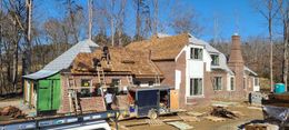 House under construction with workers, scaffolding, and a partially finished roof in a wooded area