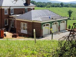 Brick house with a metal roof and white garage doors beside a green hillside and fields