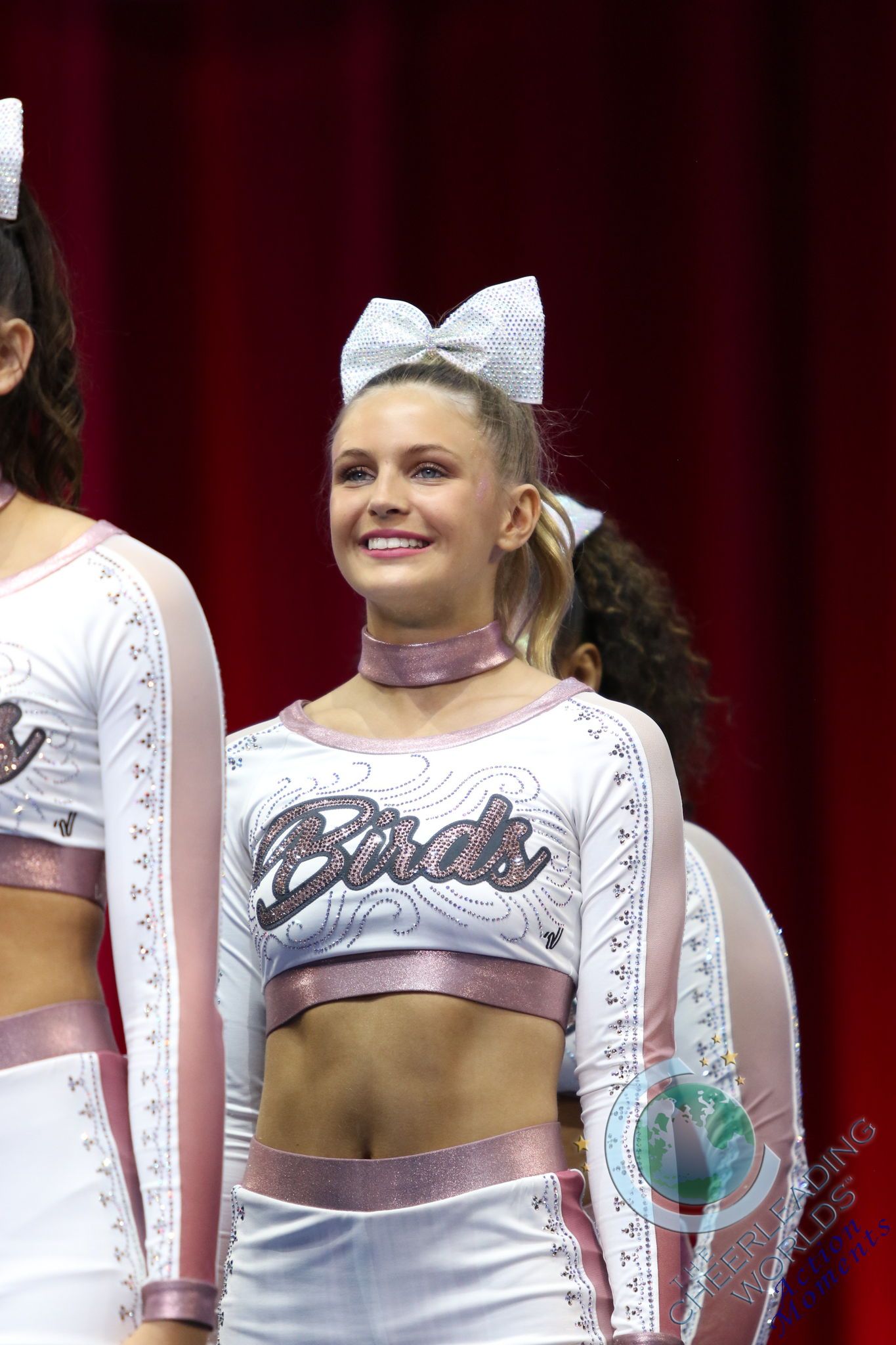 Cheerleaders in white and rose-gold uniforms with sparkling hair bows smile while standing on a stage against a red backdrop.