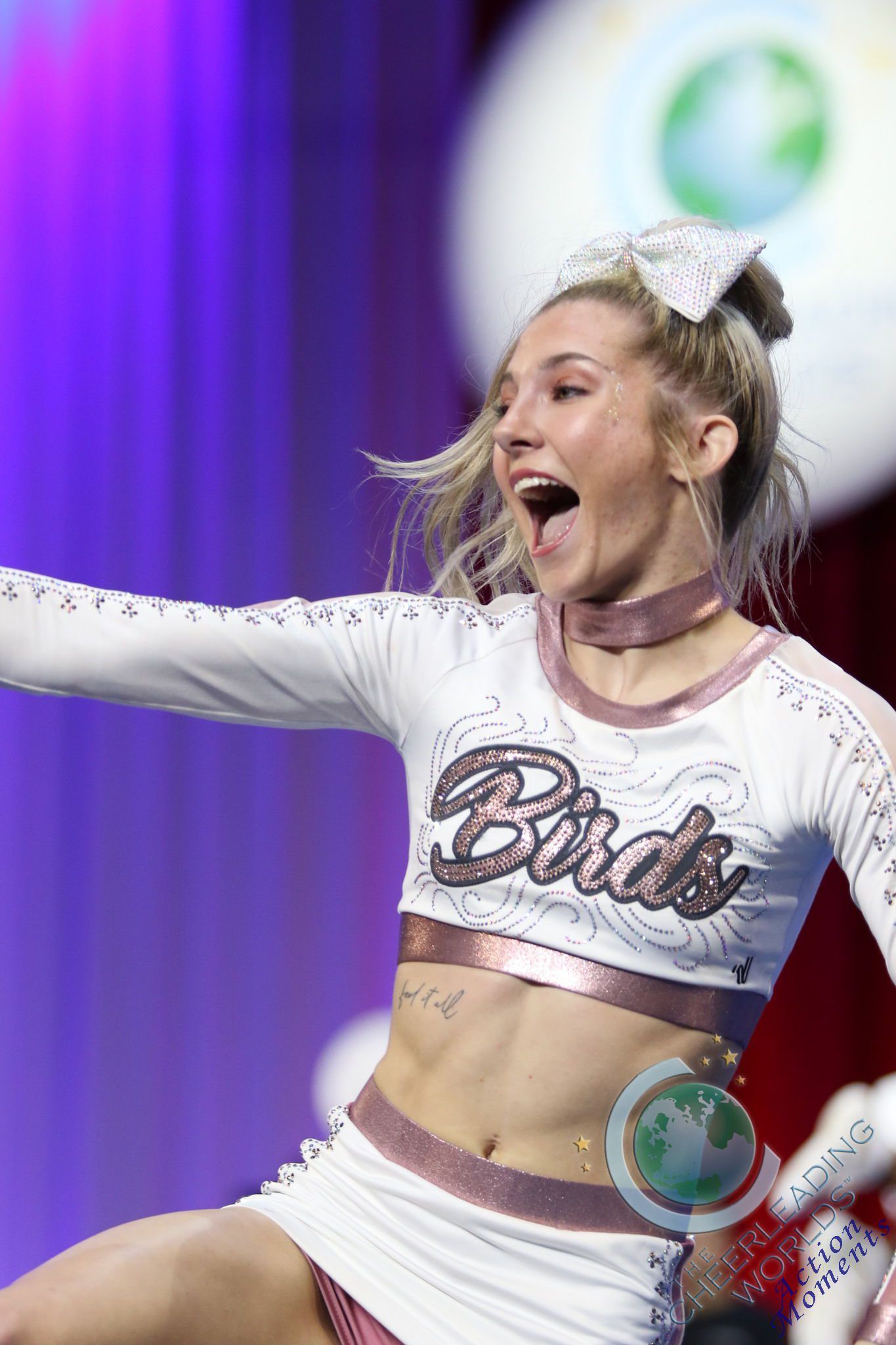A cheerleader in a white and rose-gold outfit, mid-performance with an enthusiastic expression, against a purple backdrop.