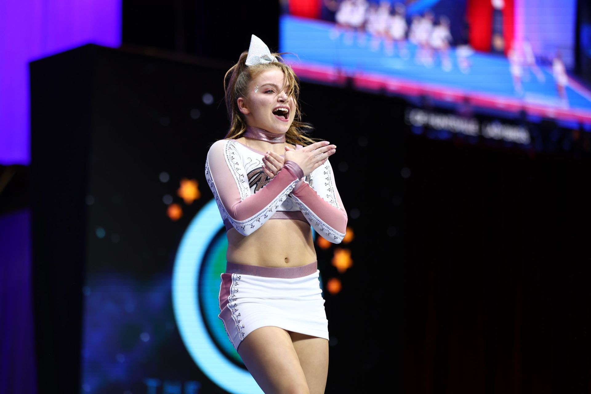 A cheerleader in a white uniform with pink accents performs on stage, arms crossed over her chest, smiling broadly.