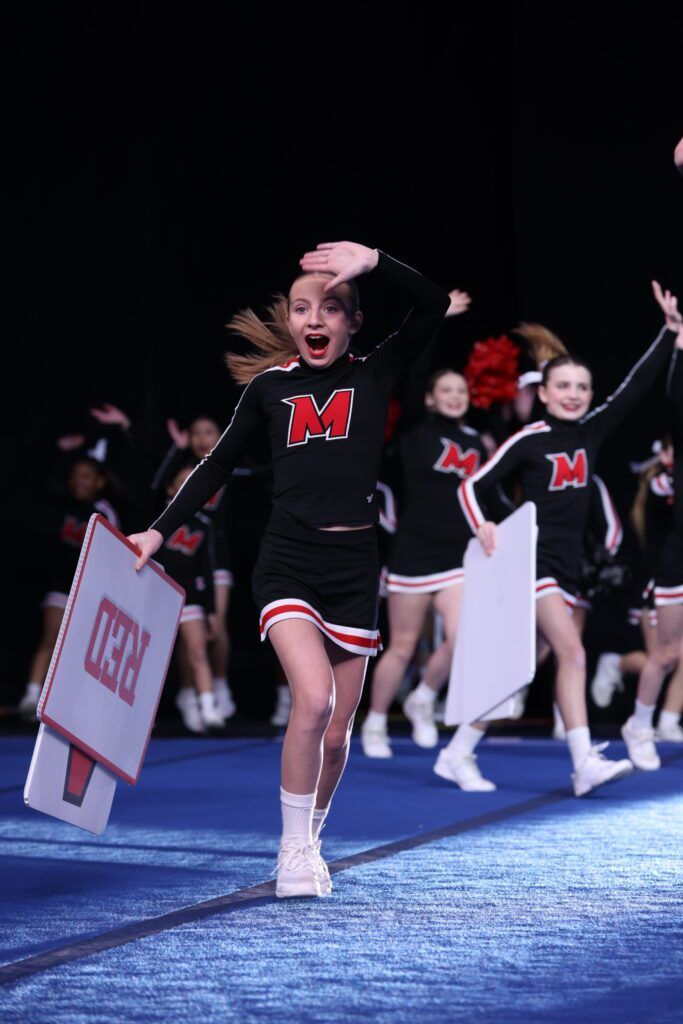 Cheerleaders in black and red uniforms perform with signs on a blue mat against a black background.