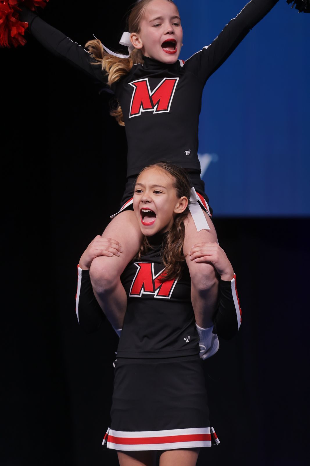 Two cheerleaders in black and red uniforms perform a stunt, both with expressions of intense focus and excitement.