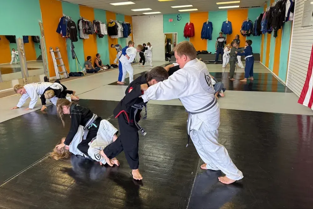 Children practicing grappling in a martial arts dojo with padded floors and hanging gear.