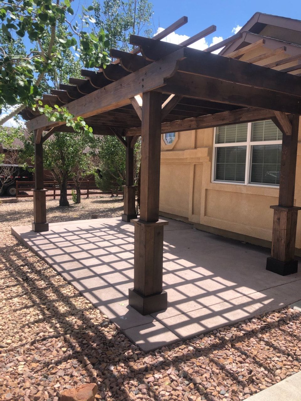 A wooden pergola over a concrete patio next to a light-colored stucco building.