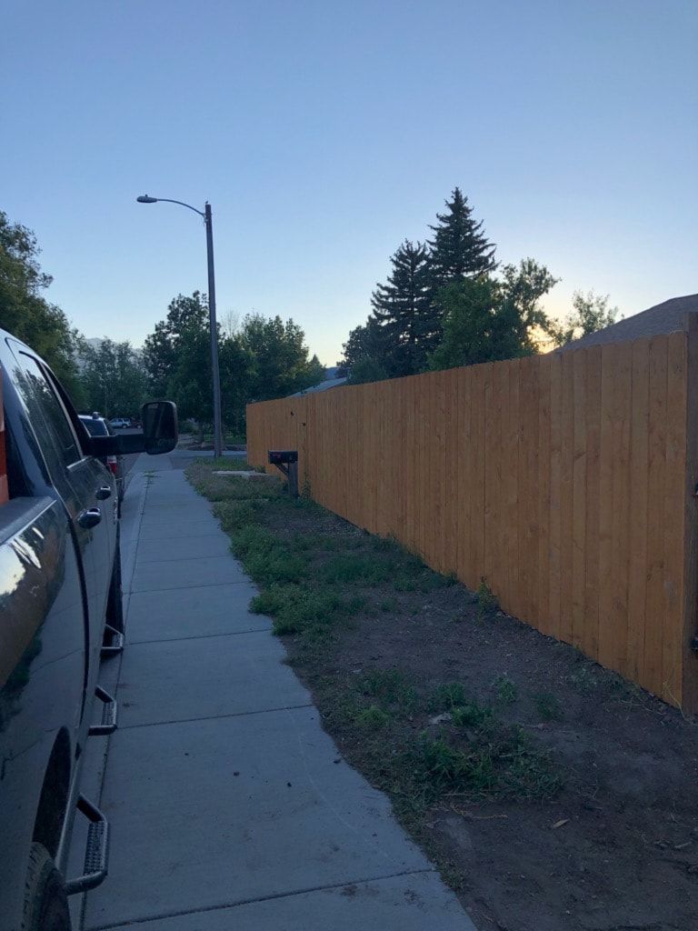 Truck parked on a sidewalk next to a wooden fence, with a street light and trees under a blue sky.