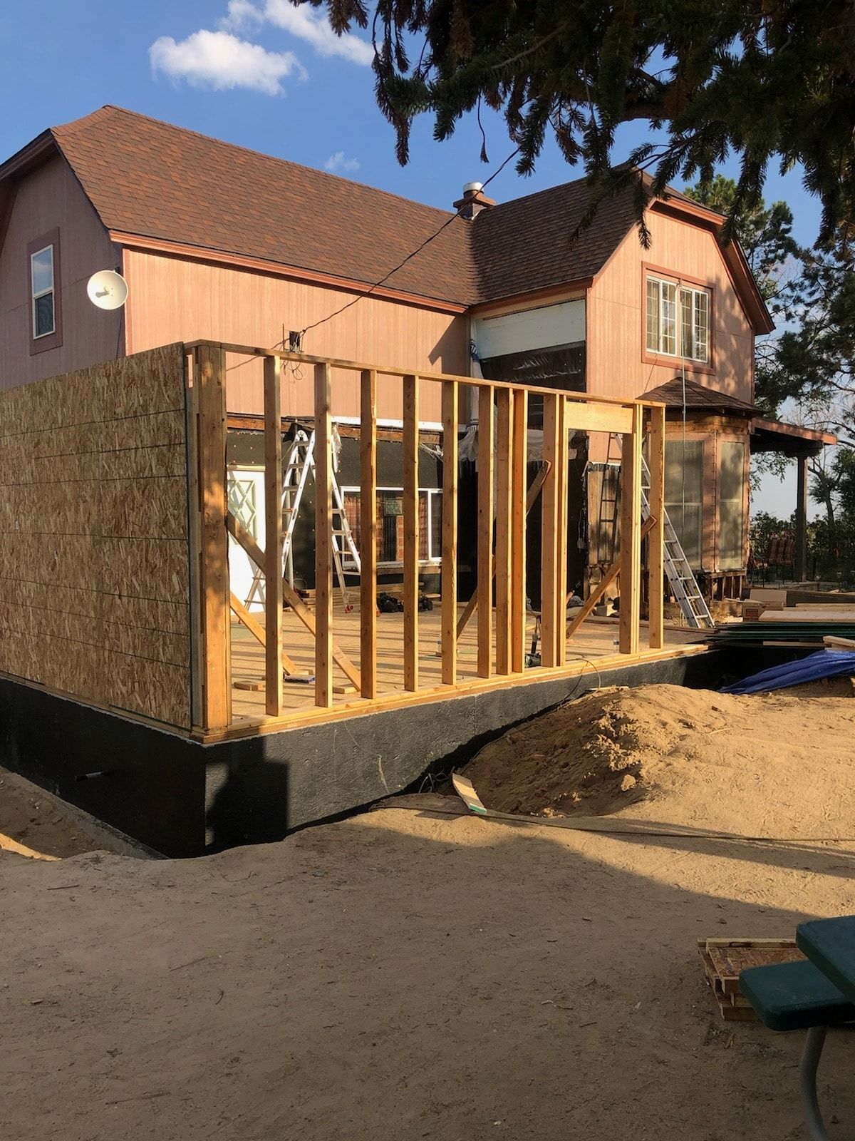 House under construction, framing a new room. Light brown wood against a pinkish-brown house. Sunny outdoor setting.