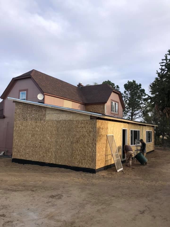 Construction of a one-story addition to a two-story house. Workers are near the windows and door, with a cloudy sky.