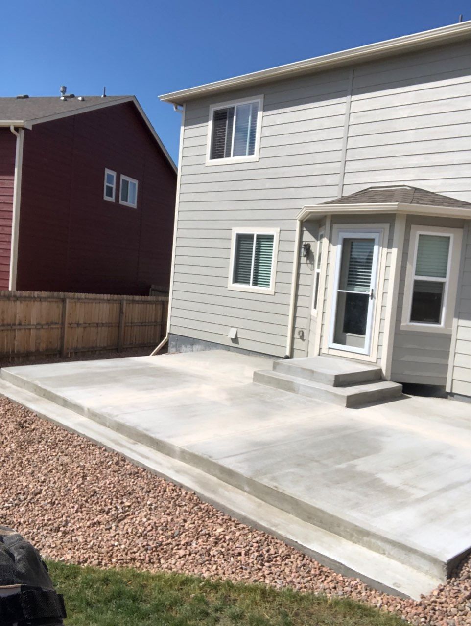 Concrete patio next to a two-story gray house. A red-brown building is to the left. Sunny day.