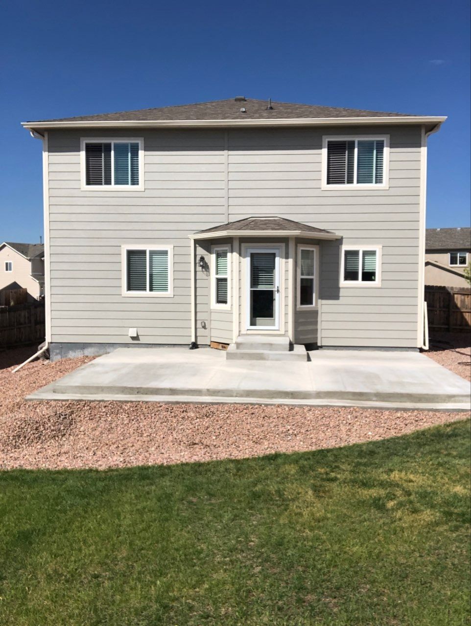 Back of a two-story gray house with a concrete patio. Red gravel surrounds the patio, green grass in foreground.