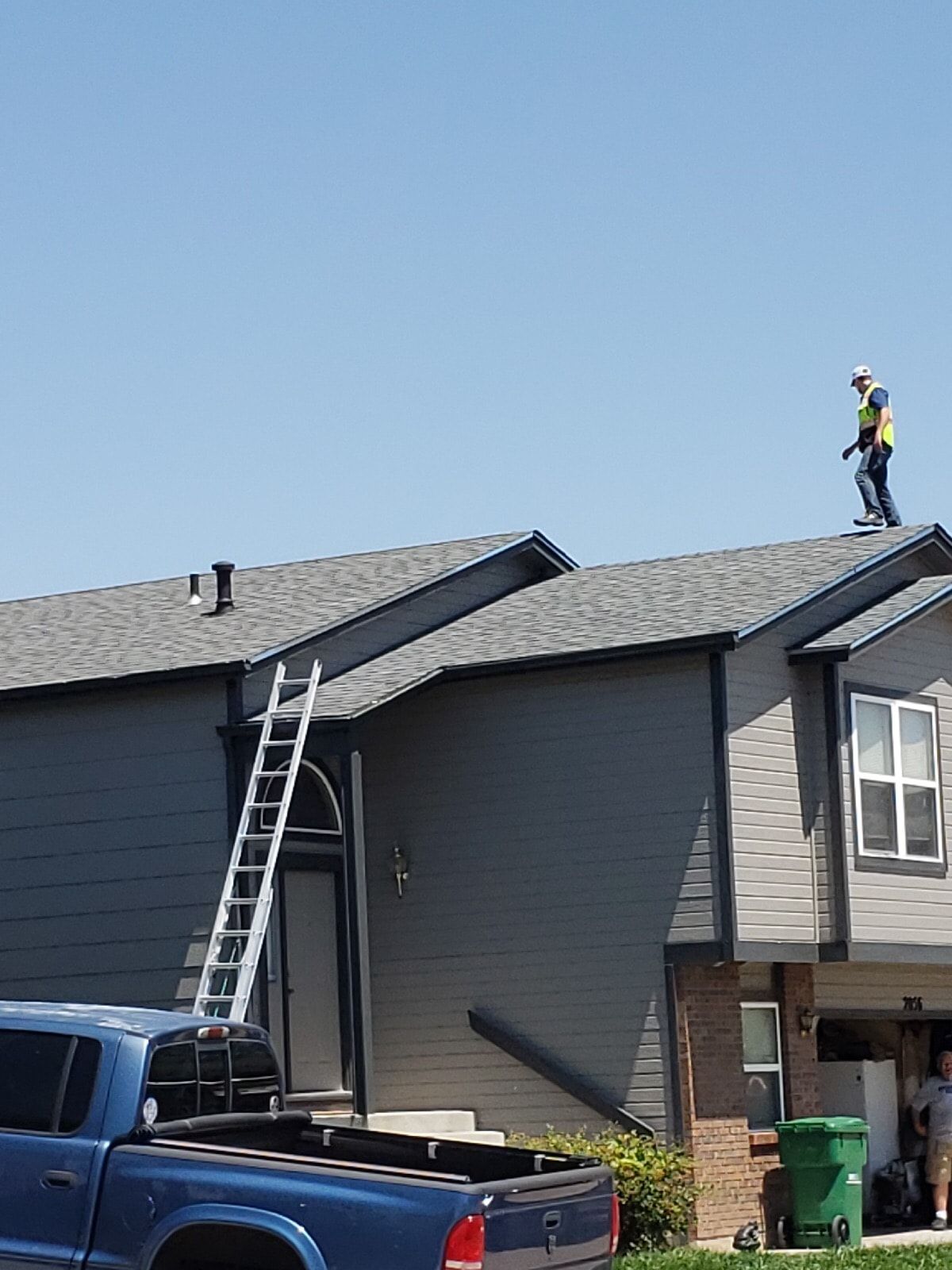 Person on a roof near a ladder, gray house and blue truck on a sunny day.