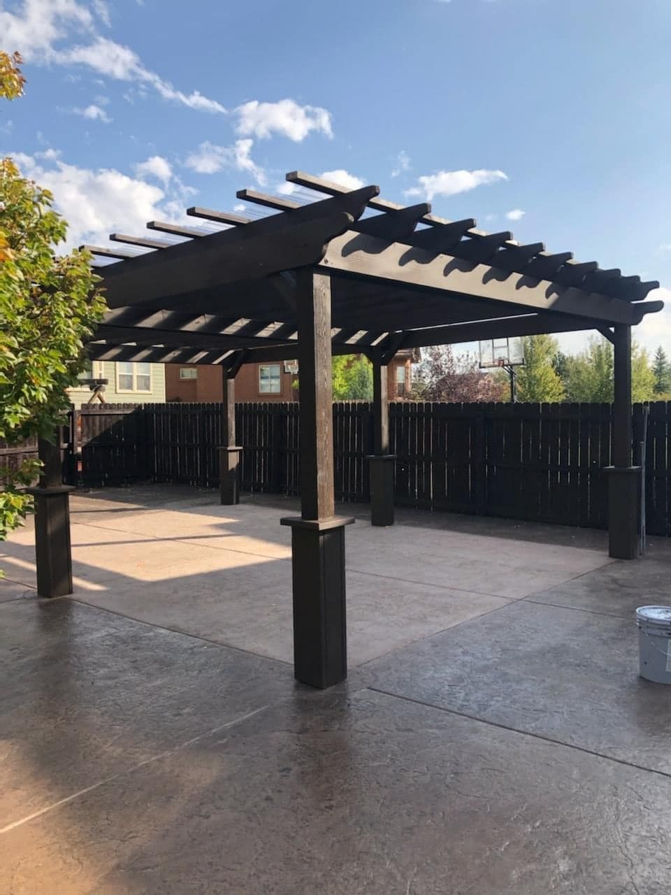 Dark wood pergola over a concrete patio. Wooden fence in background, blue sky.