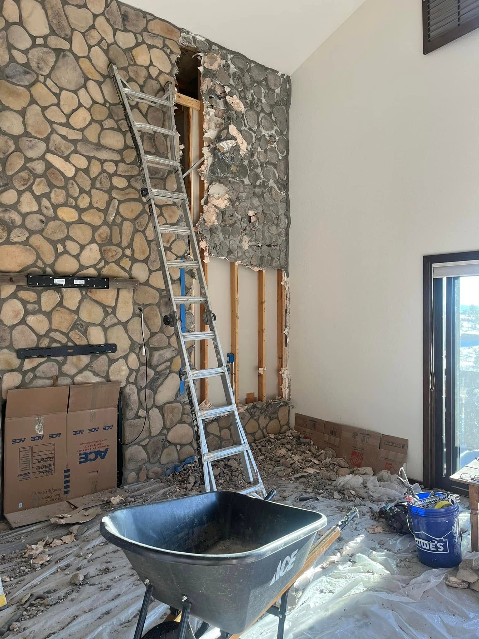 Room undergoing renovation with stone wall, ladder, wheelbarrow, and debris.
