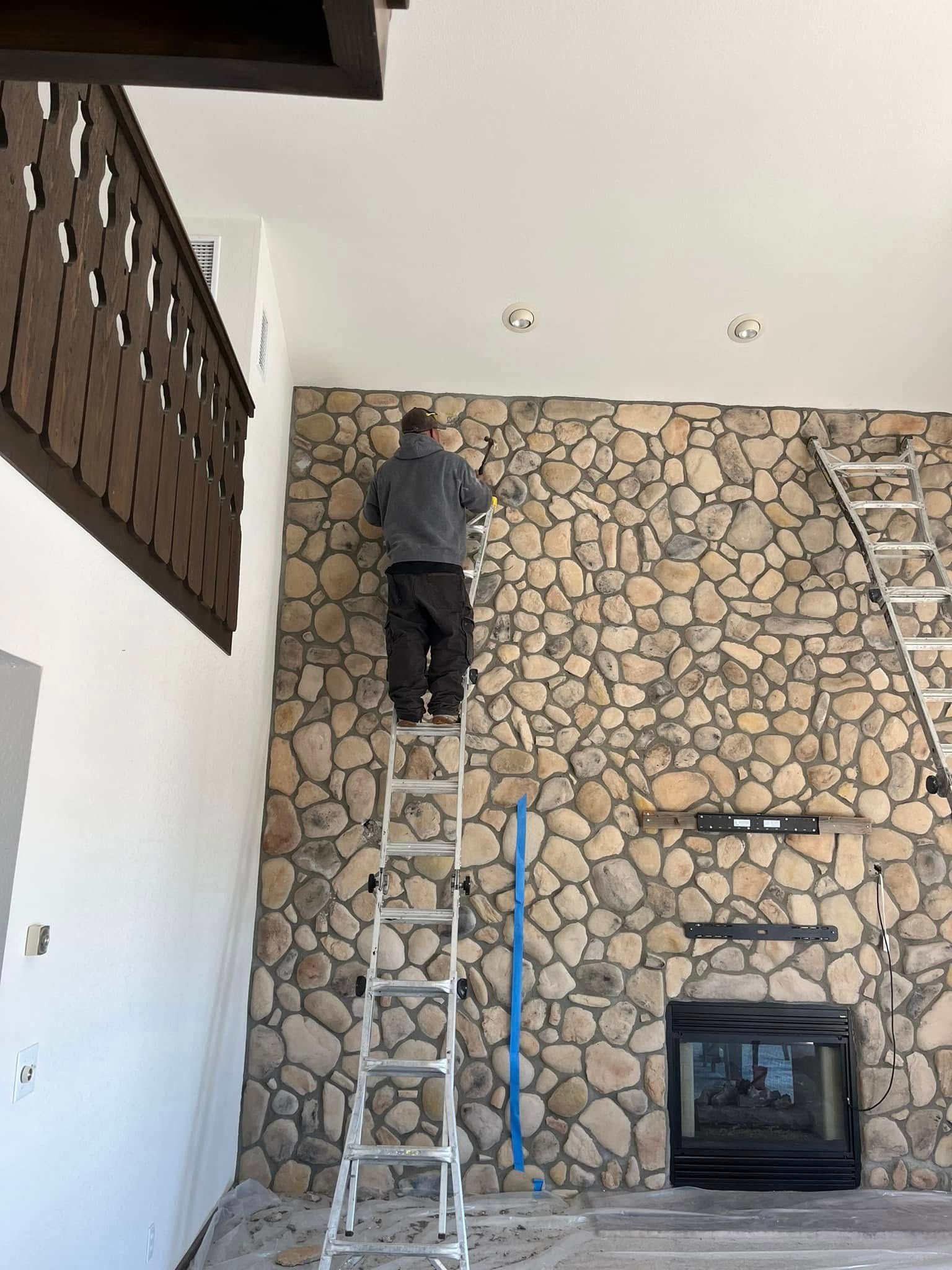 Person on ladder working on a stone wall, near a fireplace, inside a home.