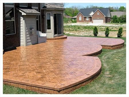 Stamped concrete patio with brick edging, tan color, near a house with a bay window.