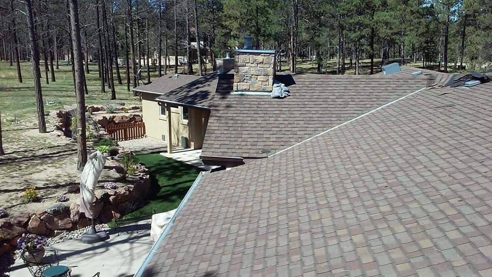 Brown shingled roof of a house with a stone chimney, surrounded by trees and a garden.