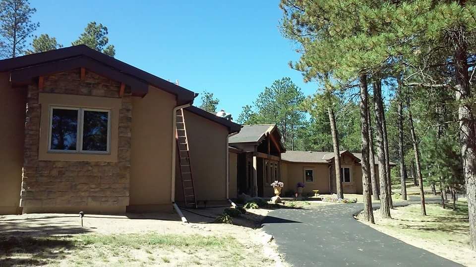 Buildings with brown stucco and stone accents under a blue sky, surrounded by pine trees.