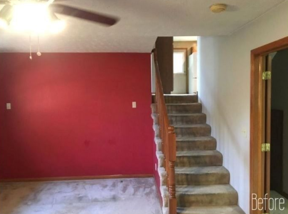Room with red accent wall, stairs leading up, and door on the right. Ceiling fan visible.