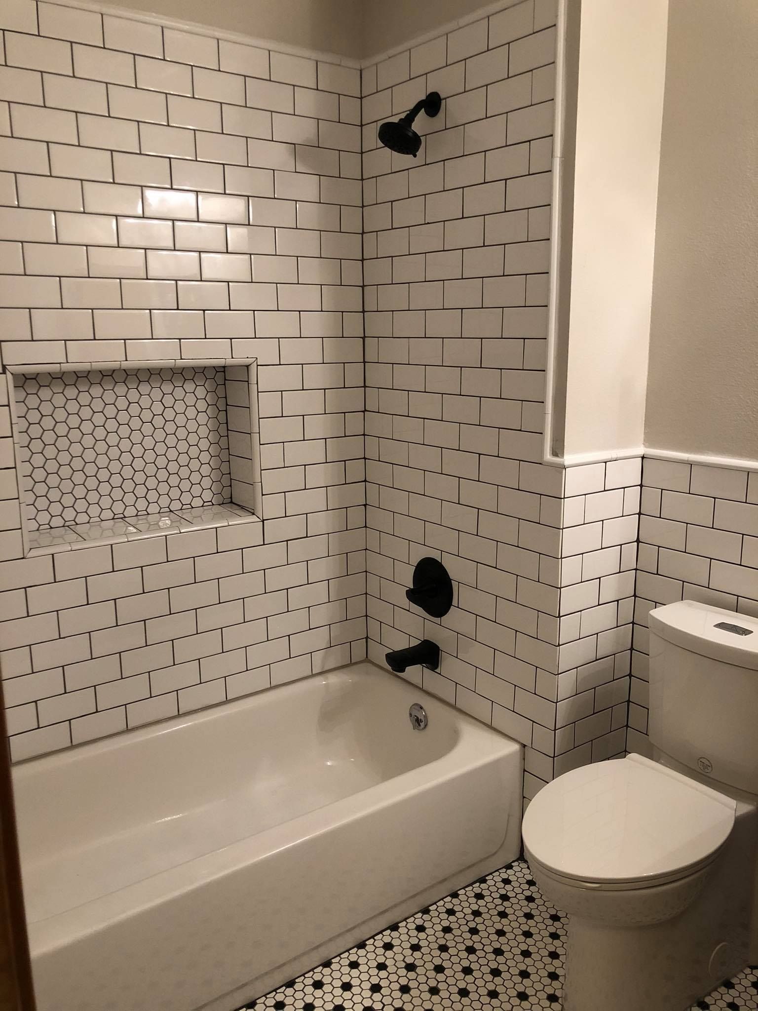 White tiled bathroom with black fixtures, shower, tub, toilet, and niche. Black and white patterned floor.