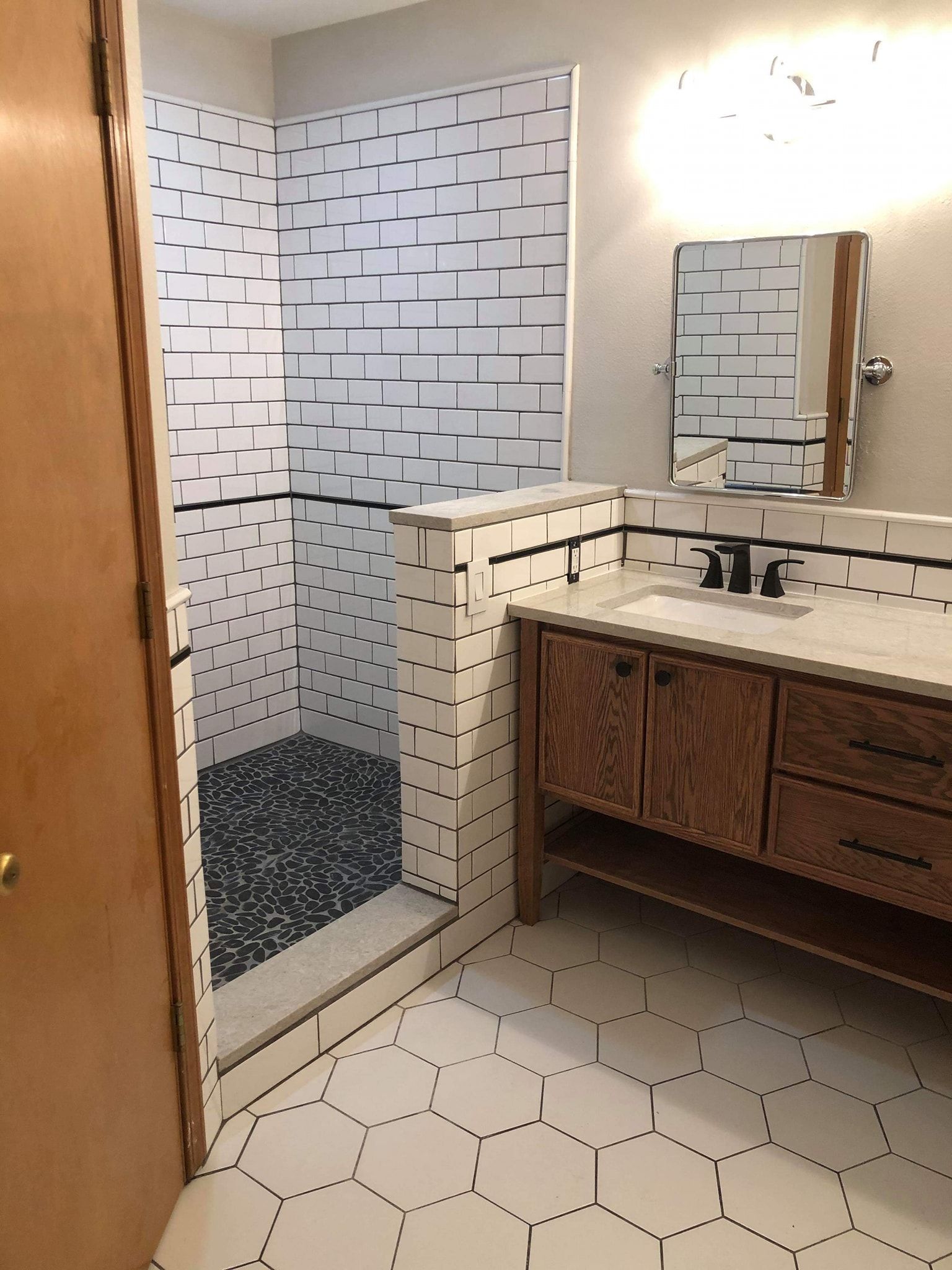 Bathroom with white hexagonal floor tiles, a shower with black and white tiles, and a wooden vanity.